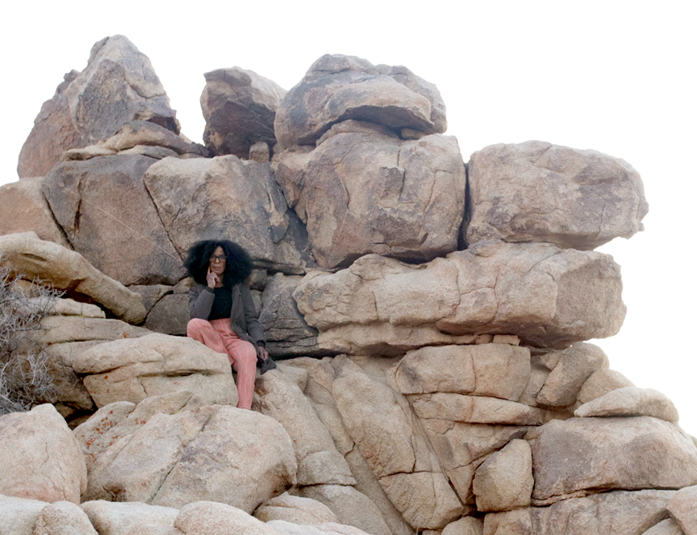 A Black femme with shoulder length natural hair and glasses sitting in a large rock formation